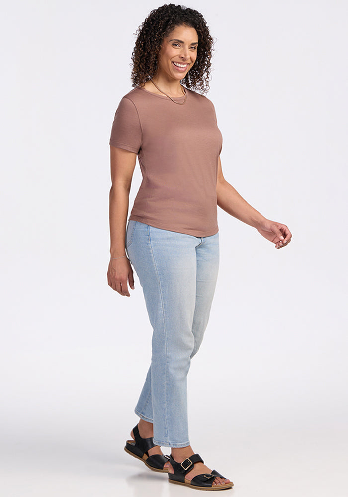 A woman with curly hair smiles as she walks, wearing the Woolx Liza Crew Neck Tee in Sienna Kiss, light blue jeans, and black sandals. The background is plain white.