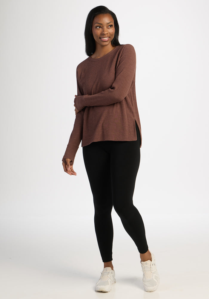 Woman in Woolx Ainsley Sweater - Sable Heather, black leggings, and white sneakers smiles against a white background.