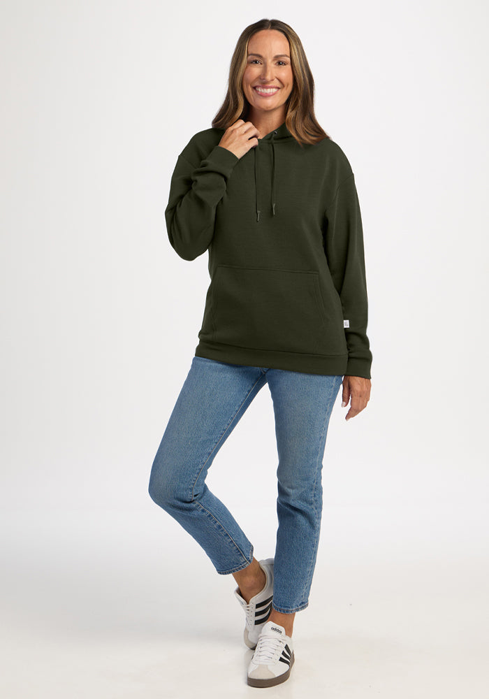 Woman in a dark green Woolx Avery Hoodie - Duffel Bag, jeans, and sneakers smiles, posing on one leg against white.