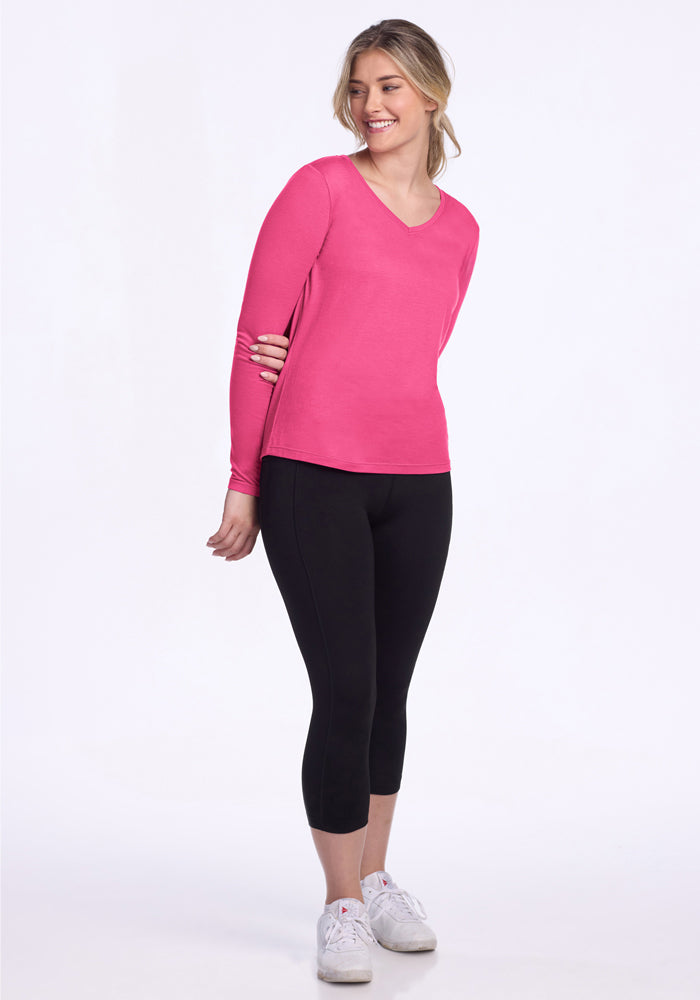 A person smiles against a white background, wearing the Woolx Millie Long Sleeve in Raspberry Fizz, black cropped leggings, and white sneakers, while holding one arm with their hand.