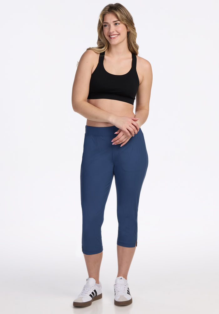 A woman smiles, wearing a black sports bra, Woolx Nicole Crop Pants in Vintage Indigo, and white sneakers with black stripes, posing against a plain white background.