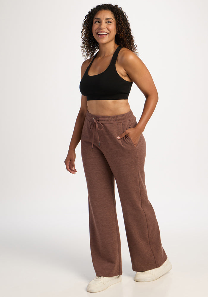 Smiling woman wears Woolx Eloise Wide Leg Pants in Sable Heather and a black sports bra, hands in pockets.