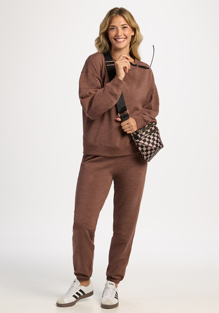 A smiling woman wears a brown Woolx Molly Crewneck - Sable Heather, matching joggers, white sneakers, and a crossbody bag.