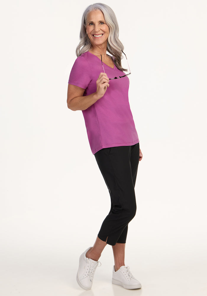 A smiling older woman with long gray hair stands sideways against a white background, wearing the Woolx Junie V Neck in Purple Wine, paired with black pants, white sneakers, and holding eyeglasses in one hand.