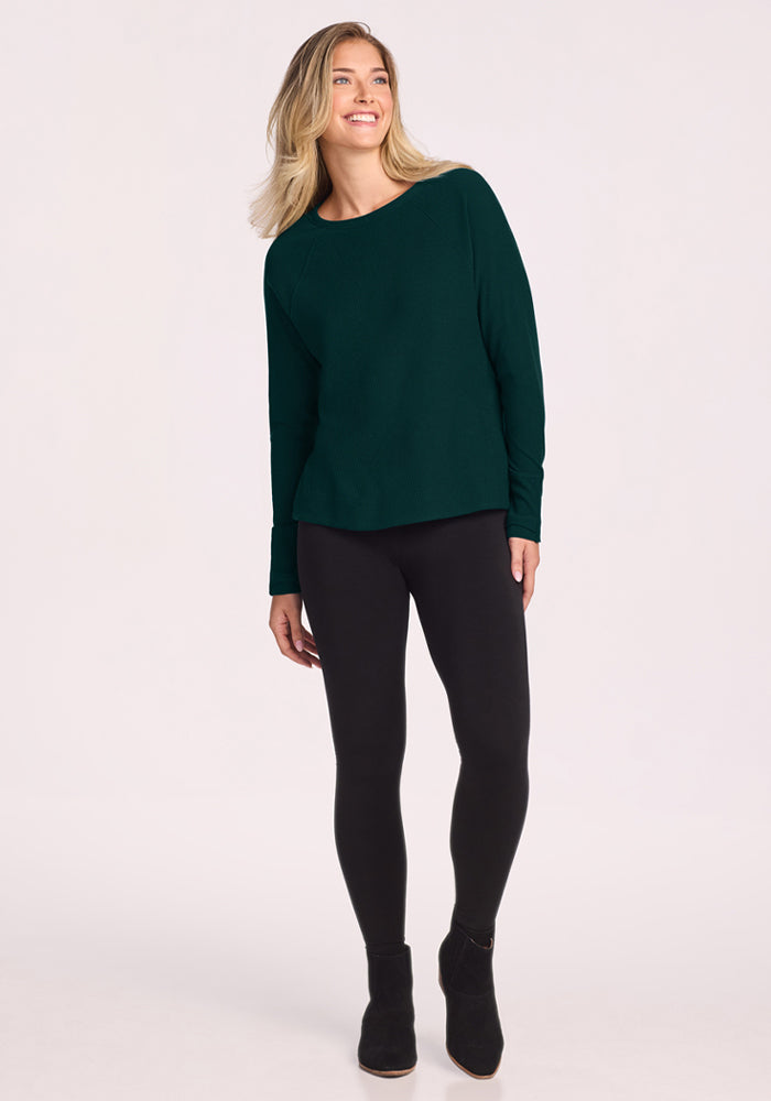 Woman in a Woolx Ainsley Sweater - Deep Pine, black leggings, and boots smiles, loving stylish temperature control.