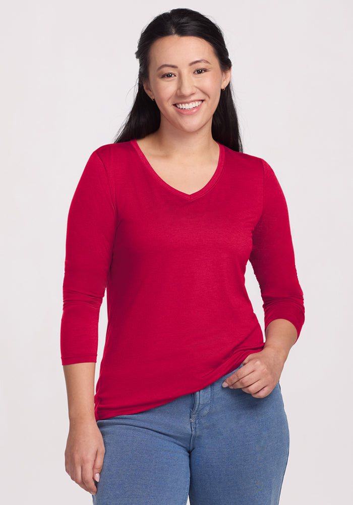 A woman with long dark hair, wearing the Woolx Millie Long Sleeve in Poppy Spark—a stylish, temperature-regulating Merino wool top—and blue jeans stands smiling with one hand in her pocket against a plain light background. 