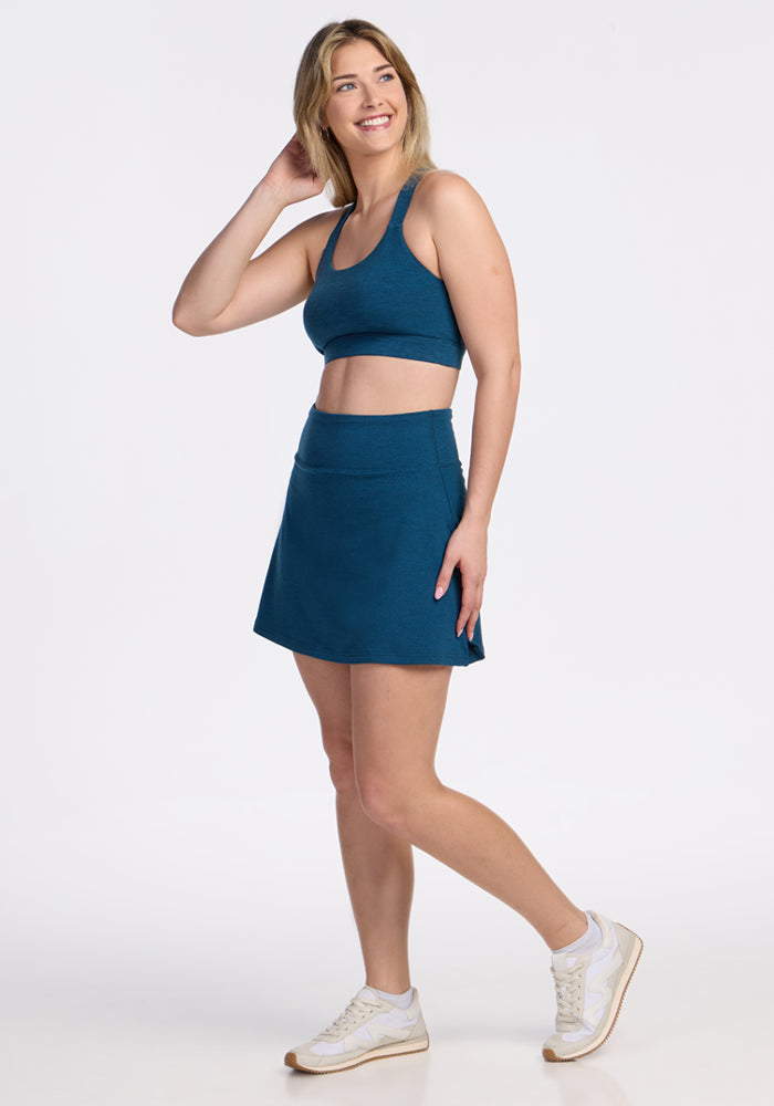 A woman smiles while posing in the Woolx Sienna Skort in Majolica Blue and a teal sports bra, paired with white sneakers, against a plain white background. 