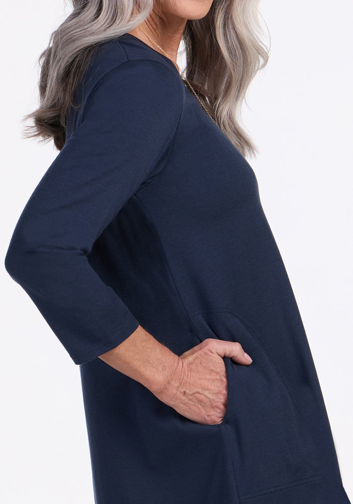 A woman with long gray hair stands sideways, right hand in a pocket, wearing the Woolx Rissa A Line Dress in Deep Navy—ideal for fall and winter layering—against a plain white background.