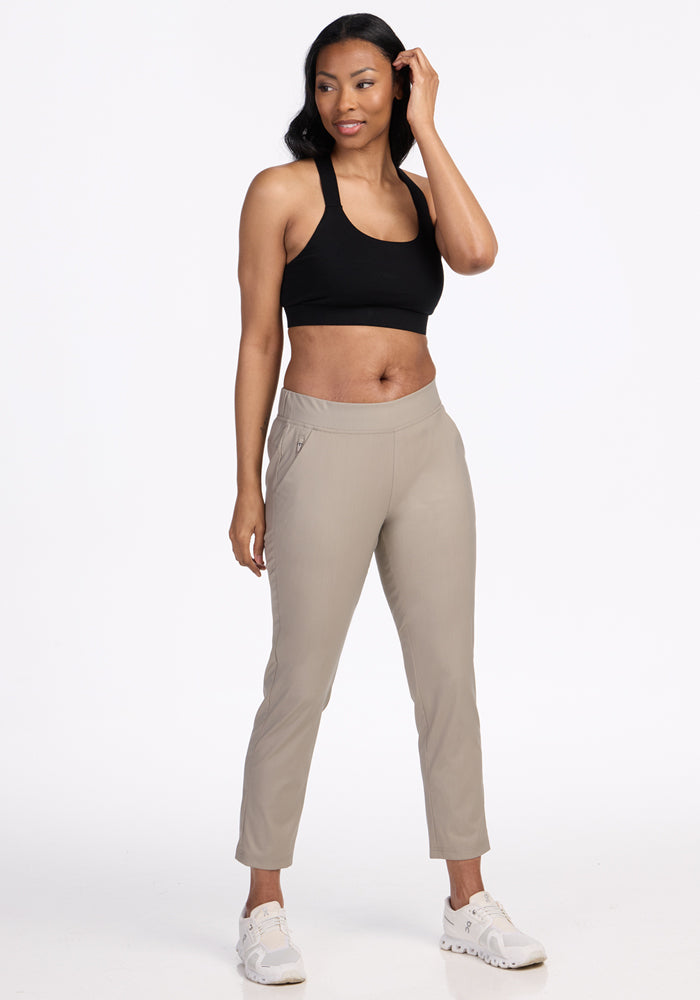 A woman smiles softly while touching her hair, wearing Woolx Nicole Ankle Pants in Vintage Khaki with a black sports bra and white athletic shoes, standing against a plain white background and looking slightly to the side.