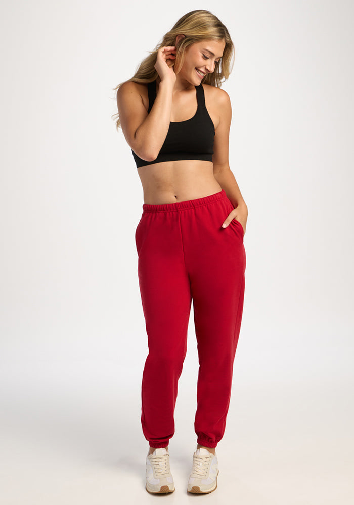 Smiling woman in Woolx Juliette Joggers - Maraschino Cherry and black sports bra stands with hand in pocket, looking down.