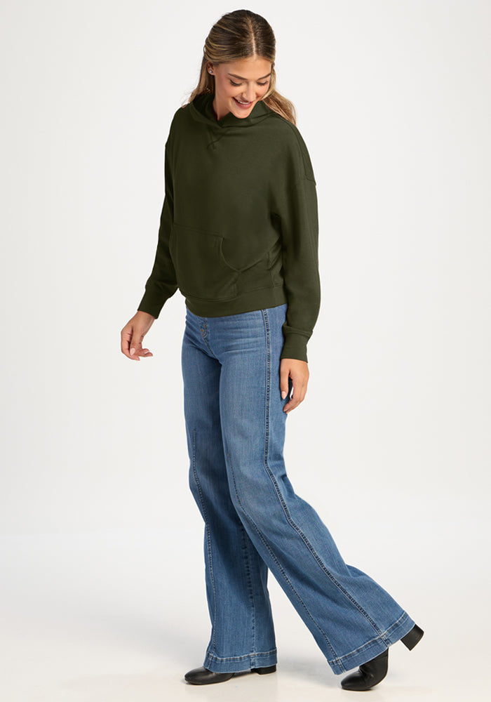 Woman in Woolx Lizzy Hoodie - Everleaf and flared jeans stands sideways, smiling, against a plain white background.