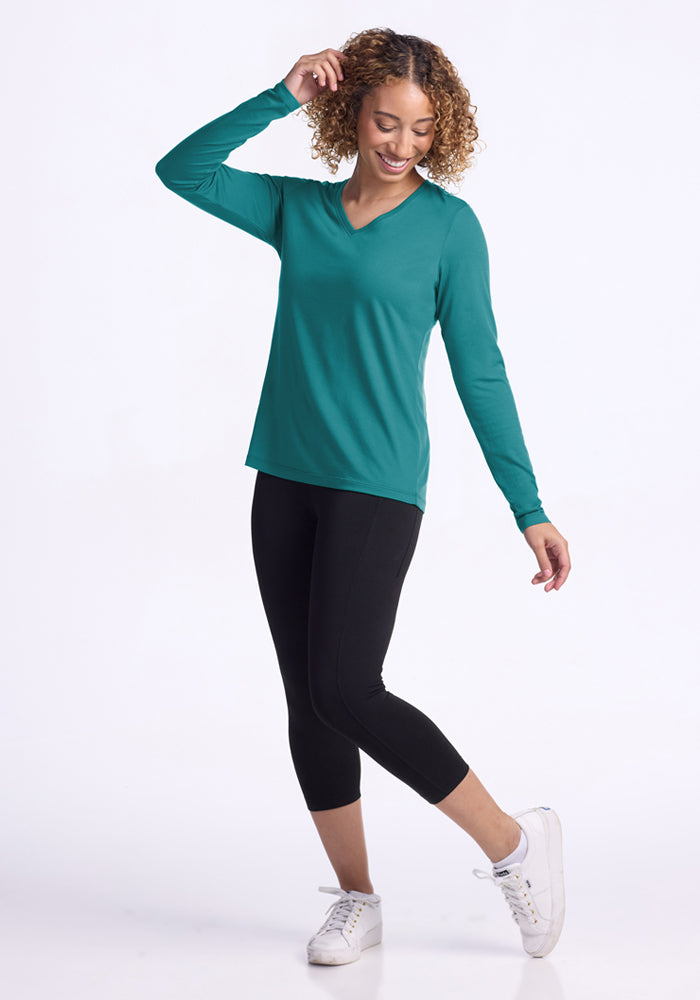 A smiling woman with curly hair wears the Woolx Millie Long Sleeve - Surfside top in teal, black cropped leggings, and white sneakers. She stands against a white background, touching her hair with one hand and relaxing the other arm.
