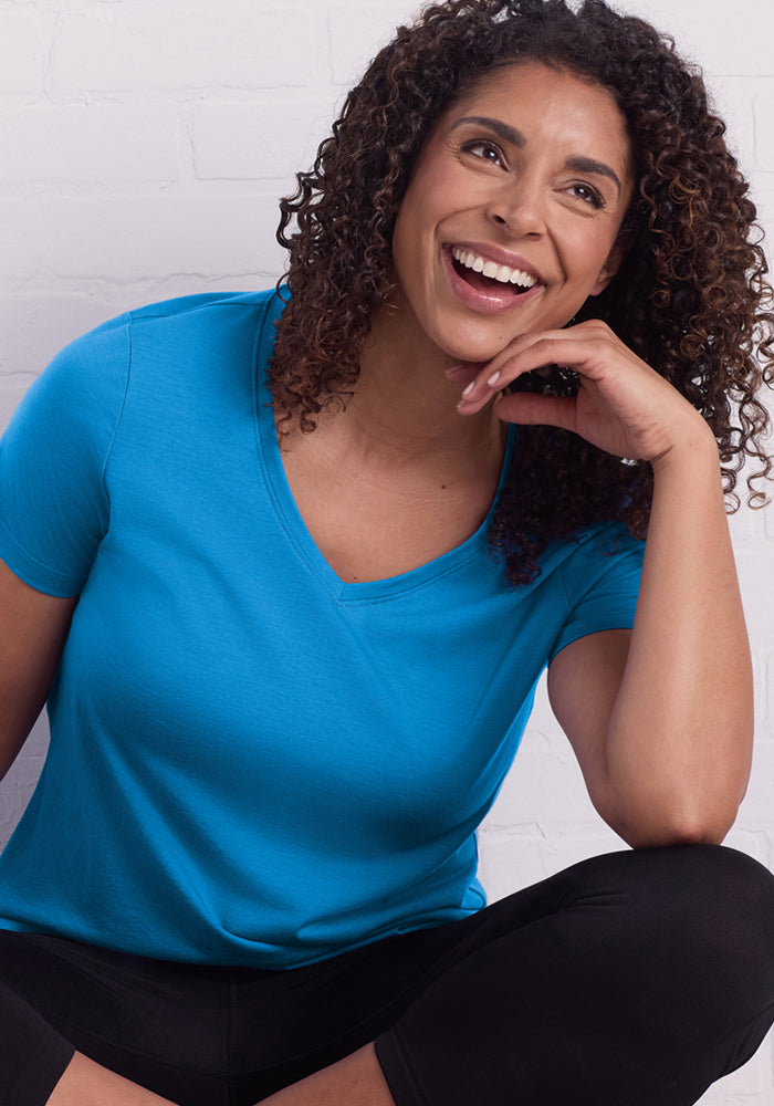 A woman with curly hair, wearing the Woolx Junie V Neck - Wave Rider women's relaxed fit Merino wool tee and black pants, smiles and looks to the side while sitting against a white brick wall.