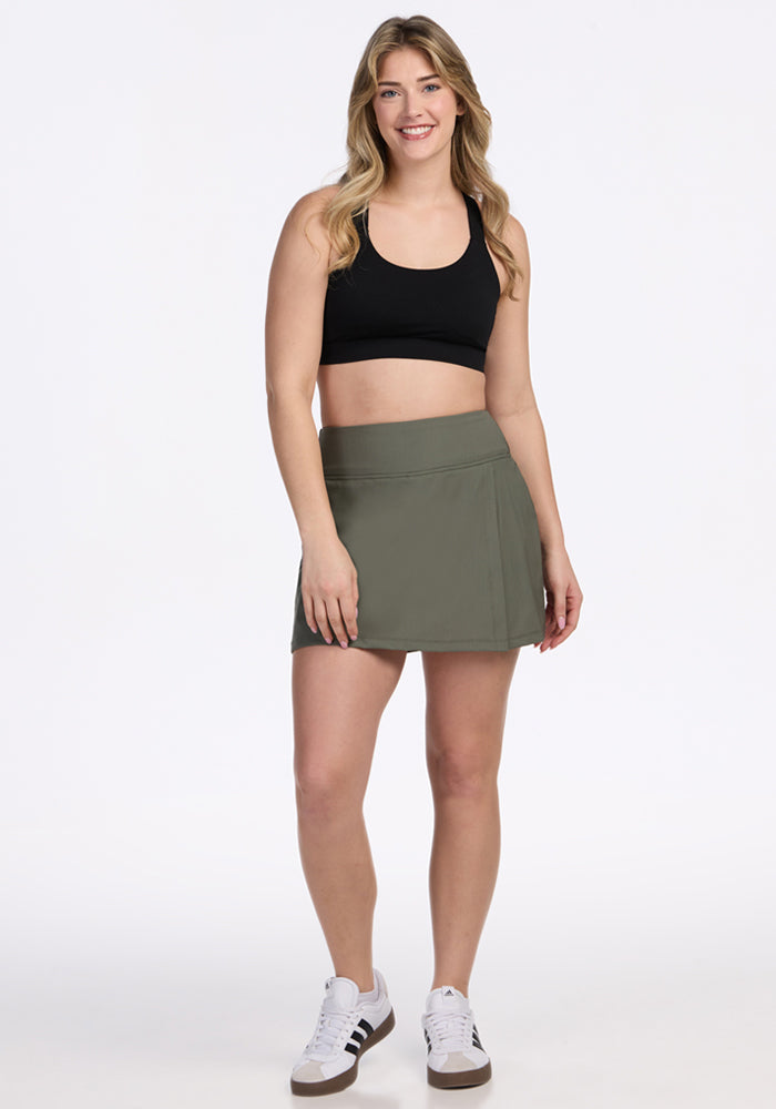 A woman smiles against a white background, wearing a black sports bra, Woolx Topanga Skort in Sage Olive with built-in shorts and pockets, and white sneakers with black stripes.