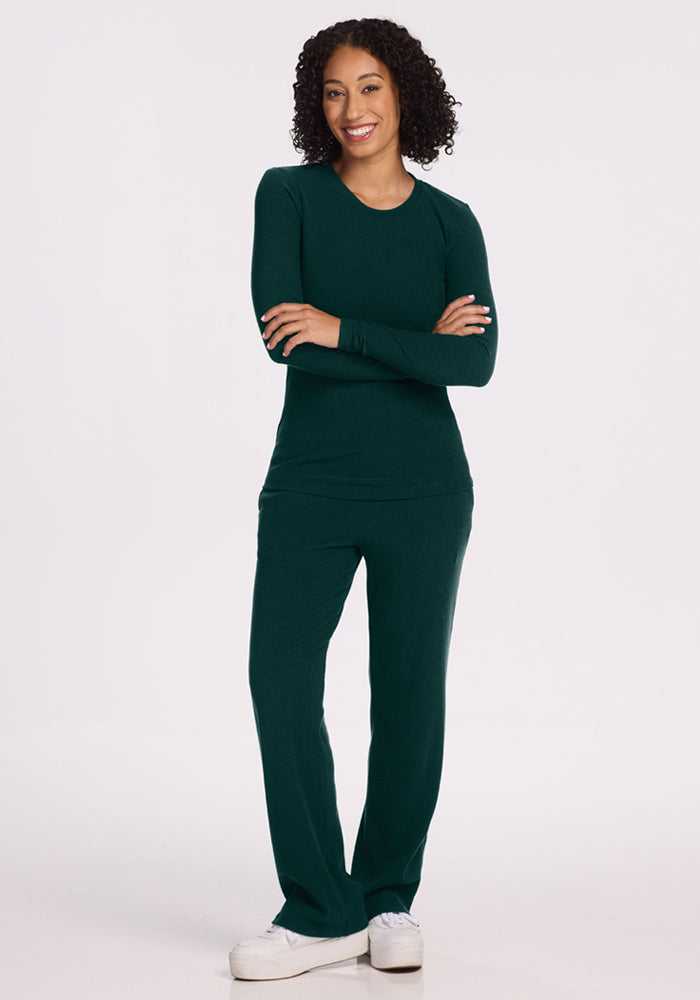 Smiling woman with curly hair wears Woolx Hadley Ribbed Crew in Deep Pine, arms crossed, on white background.
