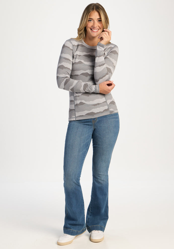 Woman in Woolx Maddie Baselayer Top - High Peaks, blue jeans, and white sneakers stands against a white background.