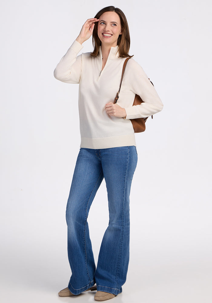 A woman with shoulder-length brown hair smiles in the Woolx Collins Half Zip Sweater in Dewdrop Pearl, paired with blue flared jeans, beige shoes, and a brown shoulder bag, posing against a plain white background.