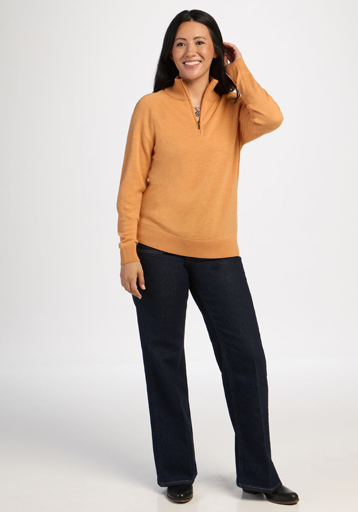 Woman in Woolx Collins Half Zip Sweater - Butterscotch and dark jeans smiles, hand in hair, on a white background.
