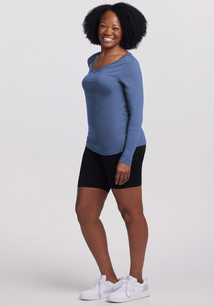 A woman with curly hair smiles while posing in the Woolx Layla V Neck Top - Skyline, paired with black shorts and white sneakers against a plain light background.