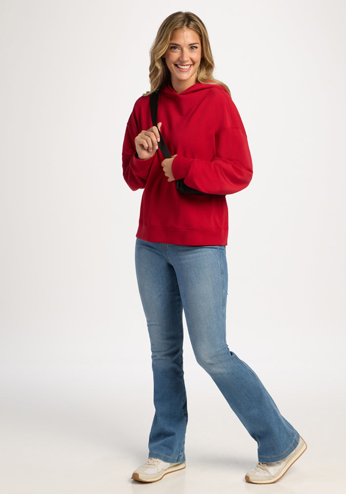 A smiling woman in a bright red Woolx Genevieve Hoodie, light blue flared jeans, and sneakers.