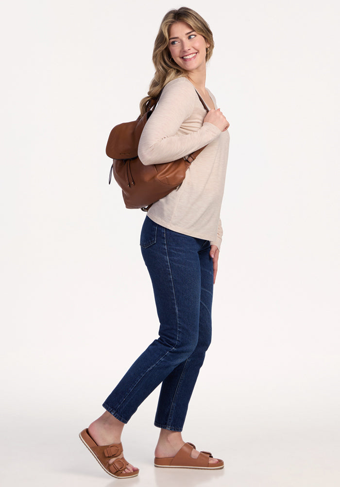 A smiling woman with long hair stands sideways against a white background, wearing the Woolx Millie Long Sleeve - Cream Heather top, paired with blue jeans, brown sandals, and a brown backpack.