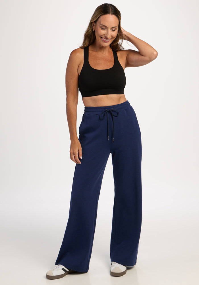 Smiling woman in black sports bra and Woolx Eloise Petite Pants - Ocean Cavern stands with one hand in her hair.