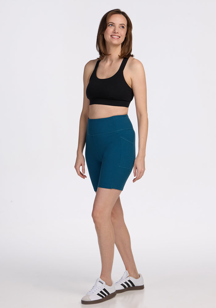 A woman with short brown hair smiles while wearing a black sports bra, Woolx Miley Bike Shorts in Moroccan Blue with pockets, and white sneakers with black stripes against a plain white background.