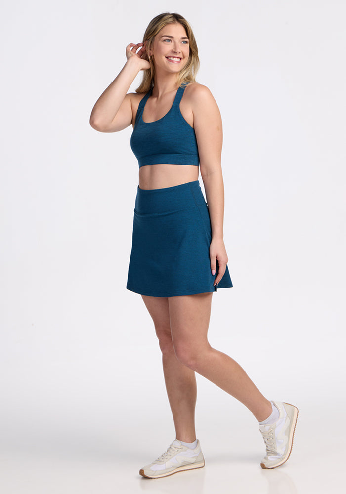A smiling woman stands on one leg against a plain white background, wearing the Woolx Livi Bra in Majolica Blue with a matching skirt and white sneakers. She touches her hair with one hand and gazes off to the side.