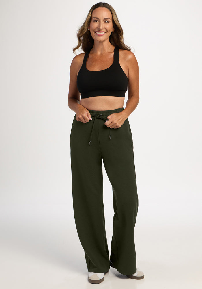 Smiling woman models Woolx Eloise Wide Leg Pants in Duffel Bag and a black sports bra against a white backdrop.