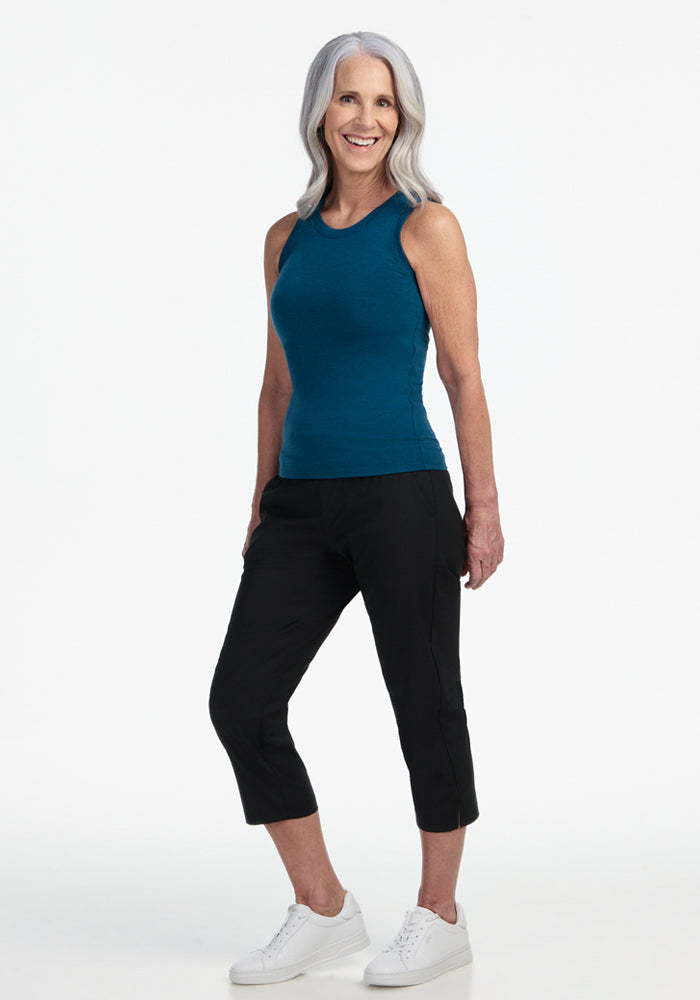 A smiling older woman with long gray hair wears the Woolx Ava Tank Top in Majolica Blue, black cropped pants, and white sneakers, standing against a plain white background.