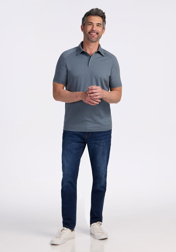 A man with short dark hair and a beard stands smiling, wearing the Woolx Summit Polo - Stormy Weather, blue jeans, and white sneakers against a plain white background.
