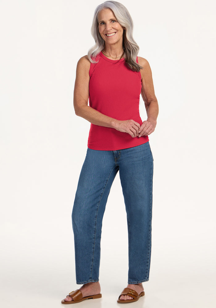 Smiling older woman wears Woolx Raegan Ribbed Tank Top in Berry Ice Pop, blue jeans, and brown sandals against plain backdrop.