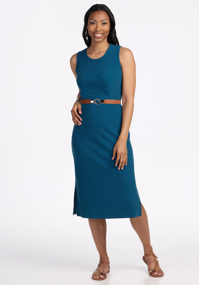A woman smiles in the Woolx Iris Ribbed Dress in Moroccan Blue, a sleeveless knee-length merino wool style with a brown belt. She wears open-toe sandals, has dark shoulder-length hair, and stands against a plain white background. 