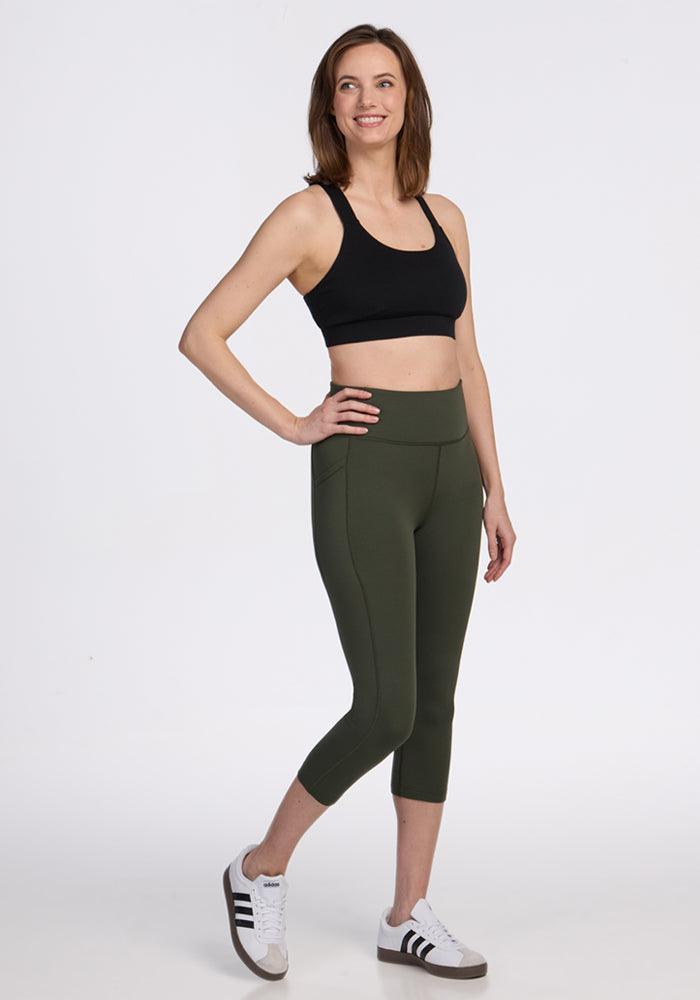 A woman stands smiling in Woolx Merino Flex® McKenna Capris - Everleaf, a black sports bra, and white Adidas sneakers against a plain white background.