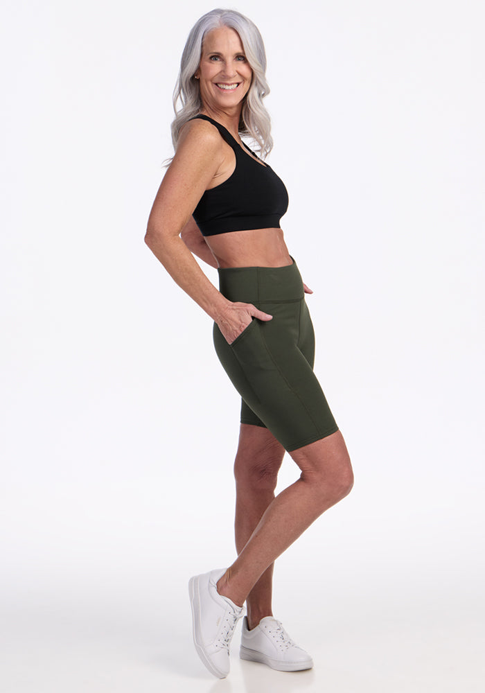 A smiling older woman with long gray hair stands in profile, wearing a black crop top, Woolx Merino Flex® Dani Bike Shorts - Everleaf with pockets, and white sneakers against a plain white background.