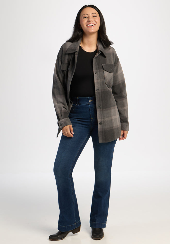 Woman smiling in the Woolx Sawyer Shacket - Volcanic Ash Plaid, black top, blue jeans, and boots on white backdrop.
