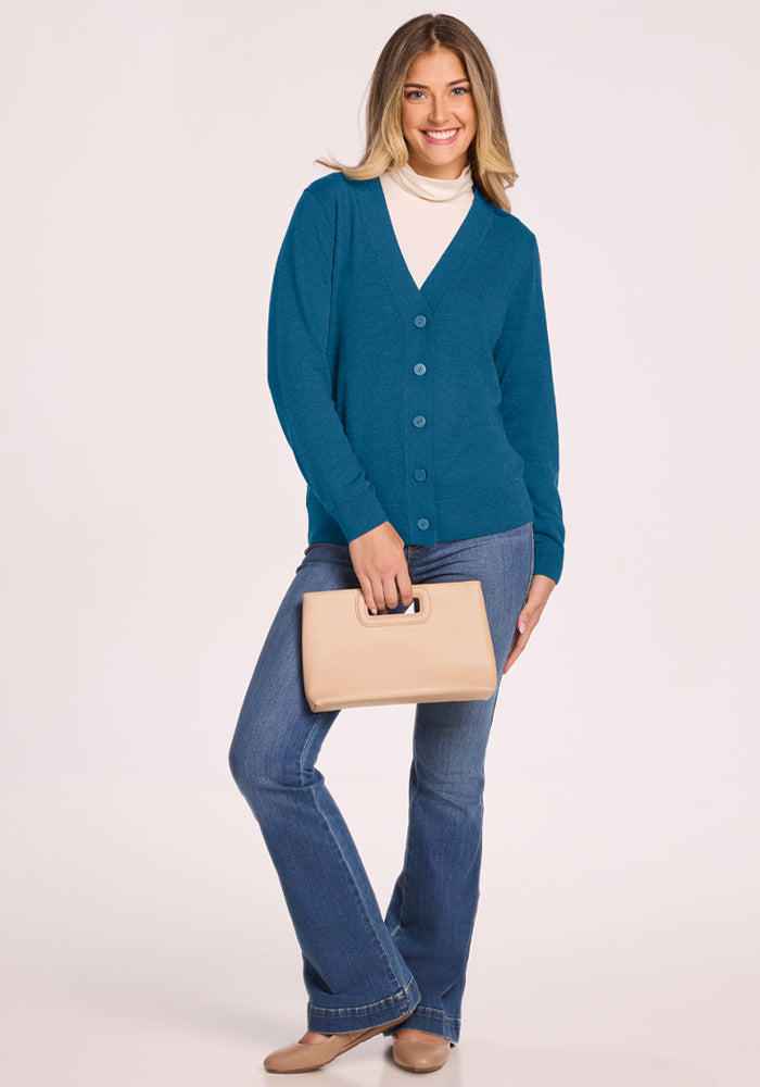 Woman smiles in Woolx Gia Sweater - Blue Pine, white top, jeans, and holds a beige handbag against a plain backdrop.