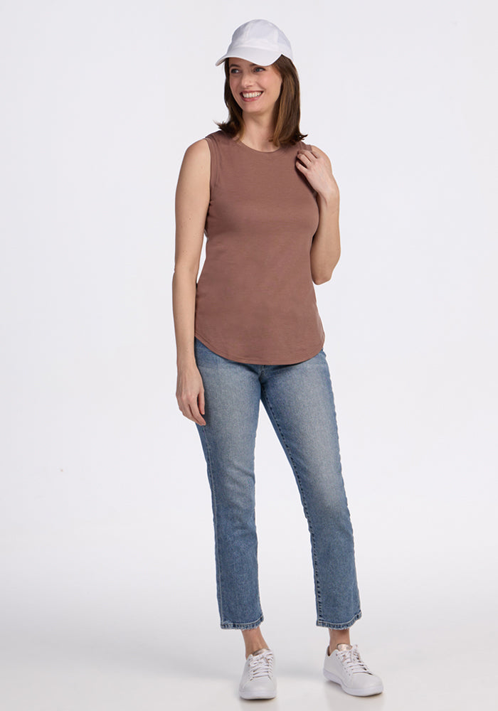 A woman smiles, wearing the Woolx Bella Tunic Top in Sienna Kiss with blue jeans, a white cap, and white sneakers against a plain white background.