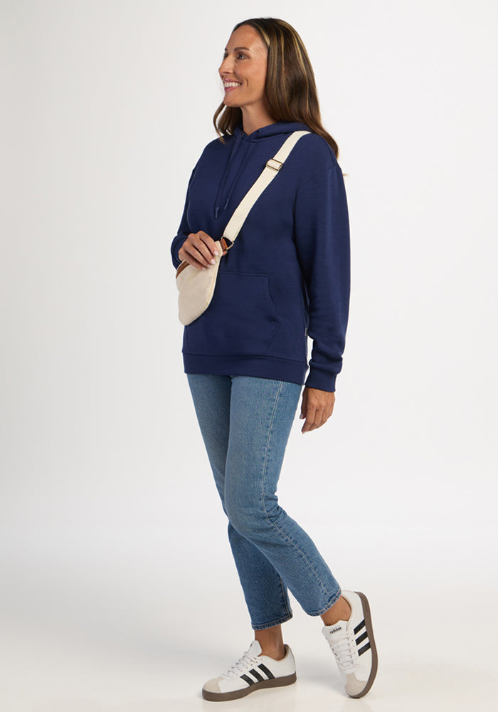 Woman in Woolx Avery Hoodie - Ocean Cavern, jeans, and sneakers smiles with a crossbody bag against a plain background.