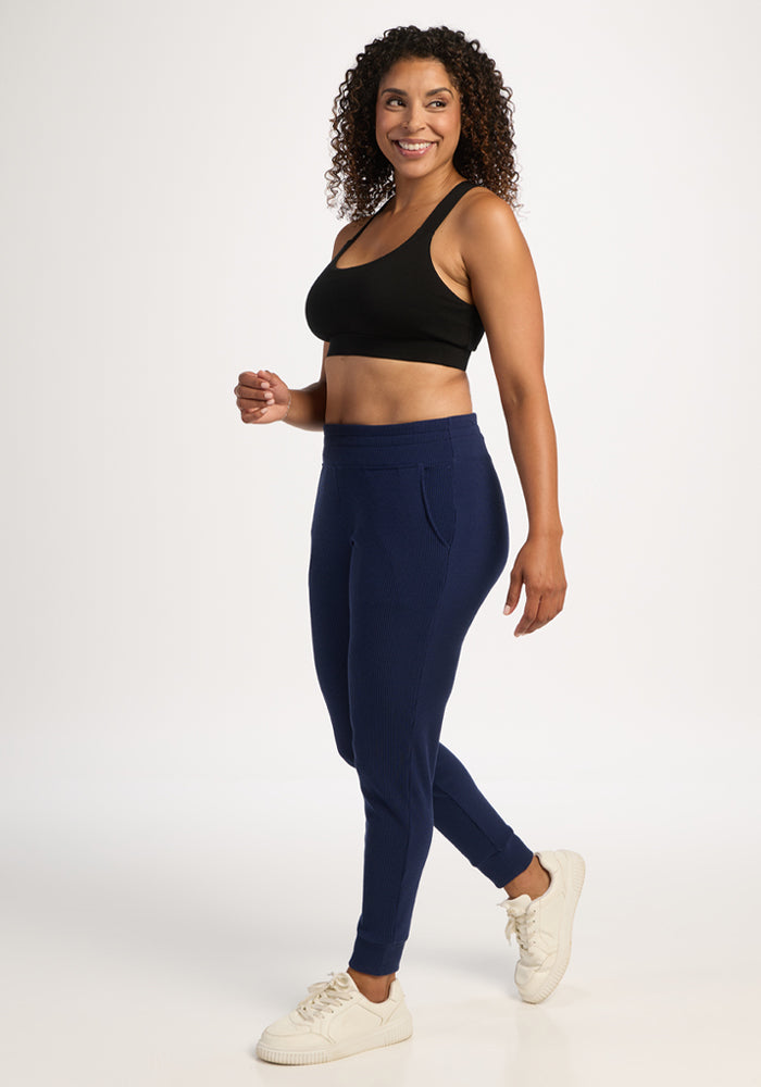 Woman in Lennon Ribbed Joggers - Ocean Cavern by Woolx walks in white sneakers, smiling against a white background.