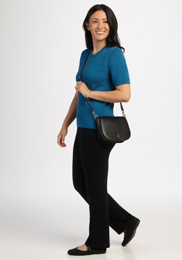 Smiling woman with long dark hair in Woolx Anna Crew Sweater - Blue Pine, black pants, flats, and a black shoulder bag.