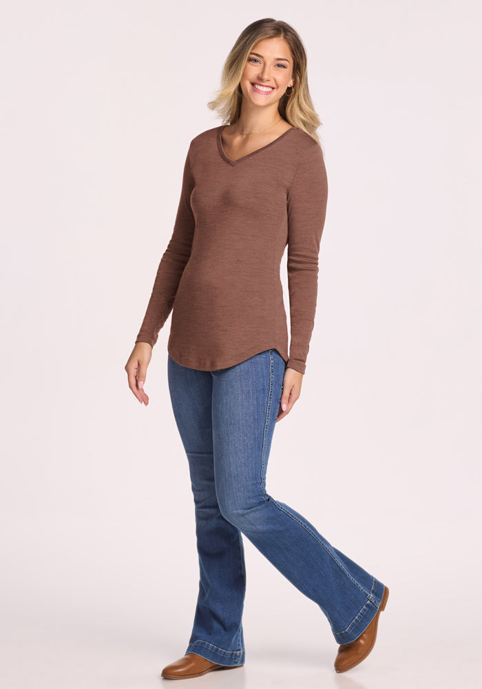 Smiling woman in Woolx Eva Tunic - Sable Heather and blue jeans poses with one foot raised against a plain background.