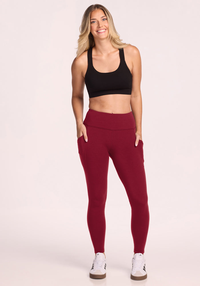 Woman in Woolx Piper Pocket Leggings - Pinot Noir and black sports bra smiles with hands in pockets against plain backdrop.