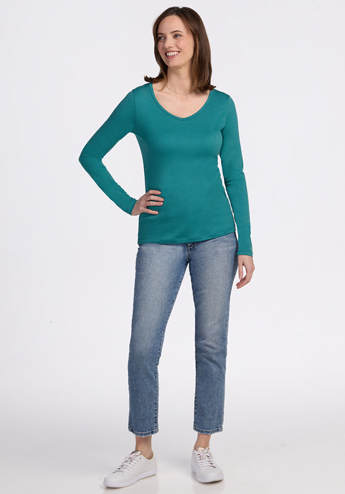 A woman smiles, posing with one hand on her hip in a Woolx Layla V Neck Top - Surfside, paired with light blue jeans and white sneakers, against a plain white background.
