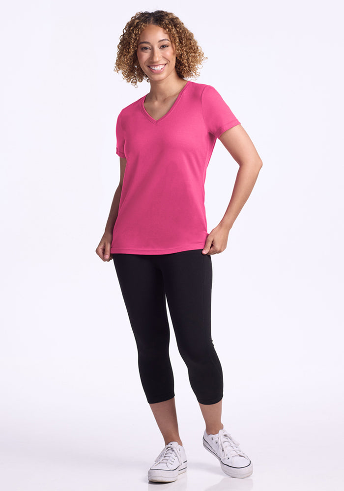 A smiling woman with curly hair wears the Woolx Junie V Neck in Raspberry Fizz, paired with black capri leggings and white sneakers. She stands against a plain white background.