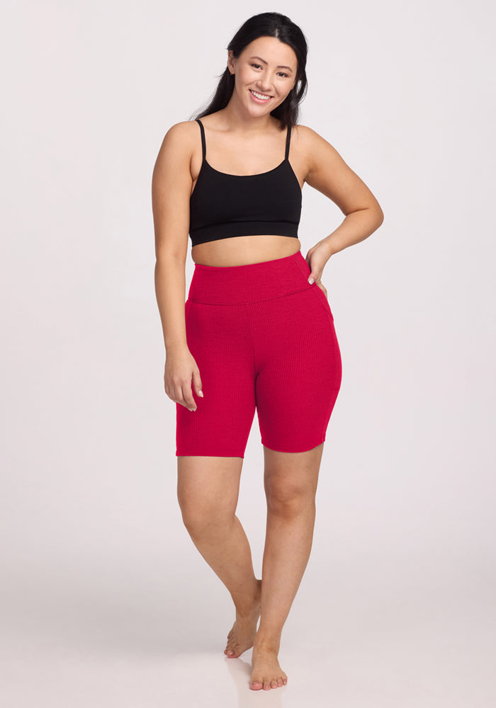 A woman stands barefoot and smiling in a black crop top and Woolx Miley Bike Shorts - Poppy Spark against a plain light background.