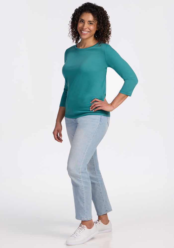 A woman with curly hair smiles while standing, wearing the Woolx Jenny 3/4 Sleeve Crew Neck - Surfside top, light blue jeans, and white sneakers against a plain white background.