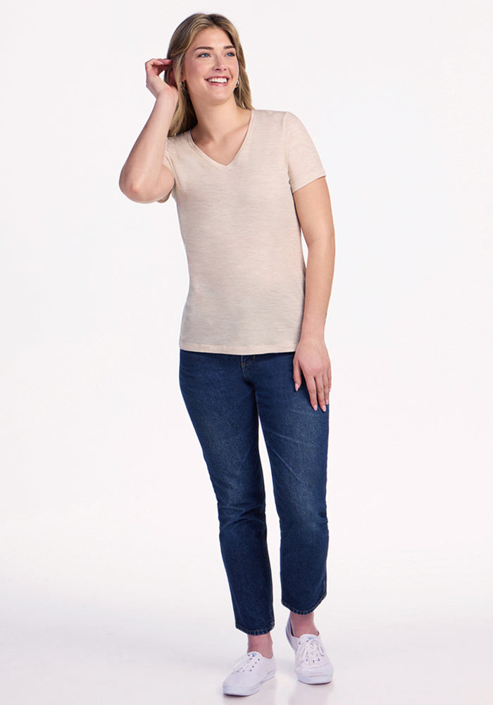 A woman with long blonde hair wears the Woolx Junie V Neck in Cream Heather, paired with blue jeans and white sneakers, smiling against a plain white background.