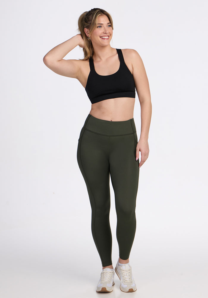 A woman smiles with one hand in her hair, wearing Woolx Merino Flex® McKenna Pocket Leggings in Everleaf, a black sports bra, and white sneakers. The background is plain white.