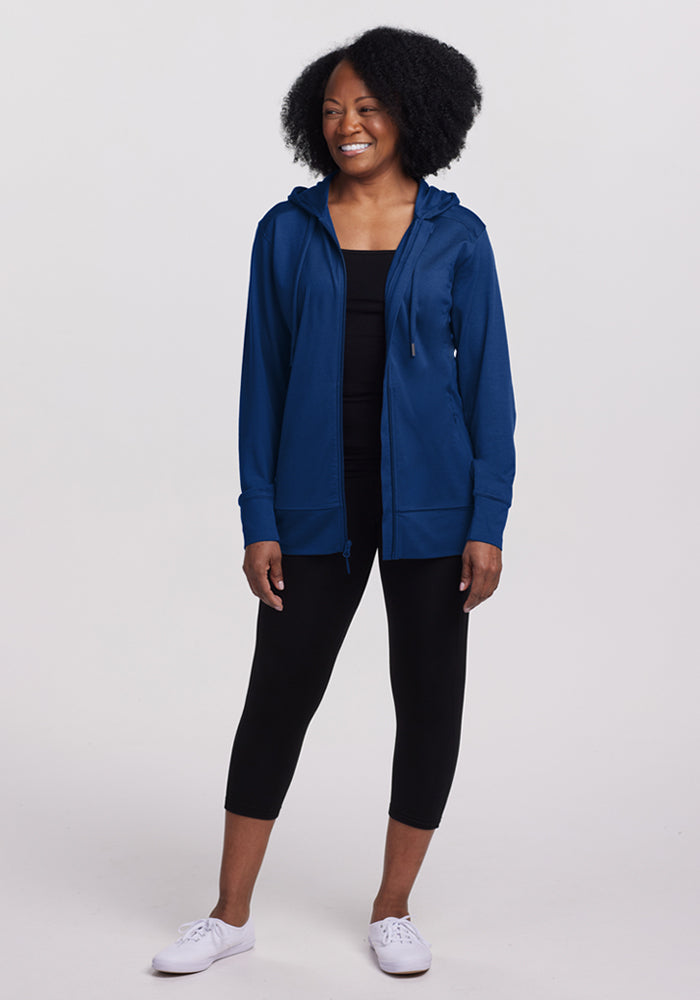 A woman with curly hair smiles, standing against a light background, wearing the Woolx Ryann Hoodie in Estate Blue over a black top with black capri leggings and white sneakers—a travel essential blending comfort and style.